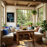 A cozy sunroom dining nook with a wooden table, cushioned bench seating, potted plants, and large windows looking out onto a lush green forest. A framed colorful koala animal print hangs on the wall.