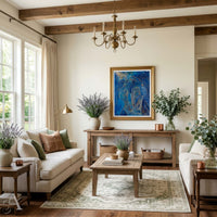 A beautifully styled living room featuring a large framed abstract blue horse painting on the wall above a wooden console table. The room is decorated with a cream sofa, a wooden coffee table on a patterned rug, potted lavender, and a rustic chandelier hanging from exposed wooden beams.