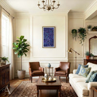 A styled living room interior with cream walls, a chandelier, two brown leather armchairs, a white sofa with blue patterned pillows, a wooden coffee table, rug, plants, and framed wall art.