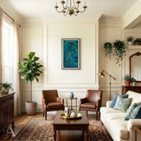 A styled living room interior with cream walls, a blue abstract wall art print, two brown leather armchairs, a white sofa with blue patterned pillows, a wooden coffee table, and multiple potted plants.